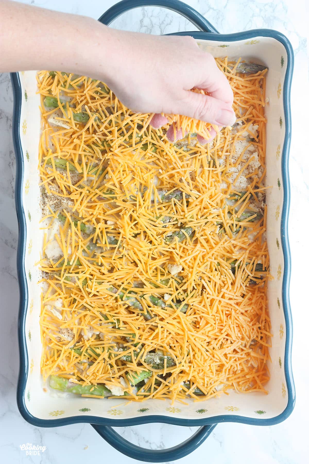 hand sprinkling cheddar cheese over the top of an asparagus casserole