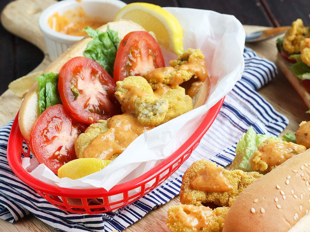 Dressed fried oyster po' boy sandwich wrapped in paper in a red plastic basket with a blue and white napkin underneath