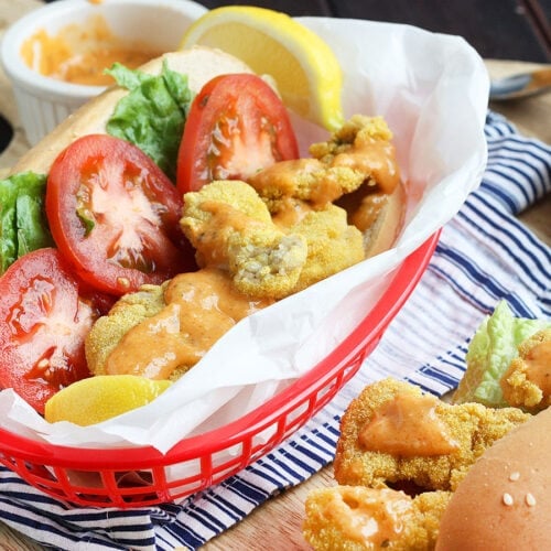 Dressed fried oyster po' boy sandwich wrapped in paper in a red plastic basket with a blue and white napkin underneath