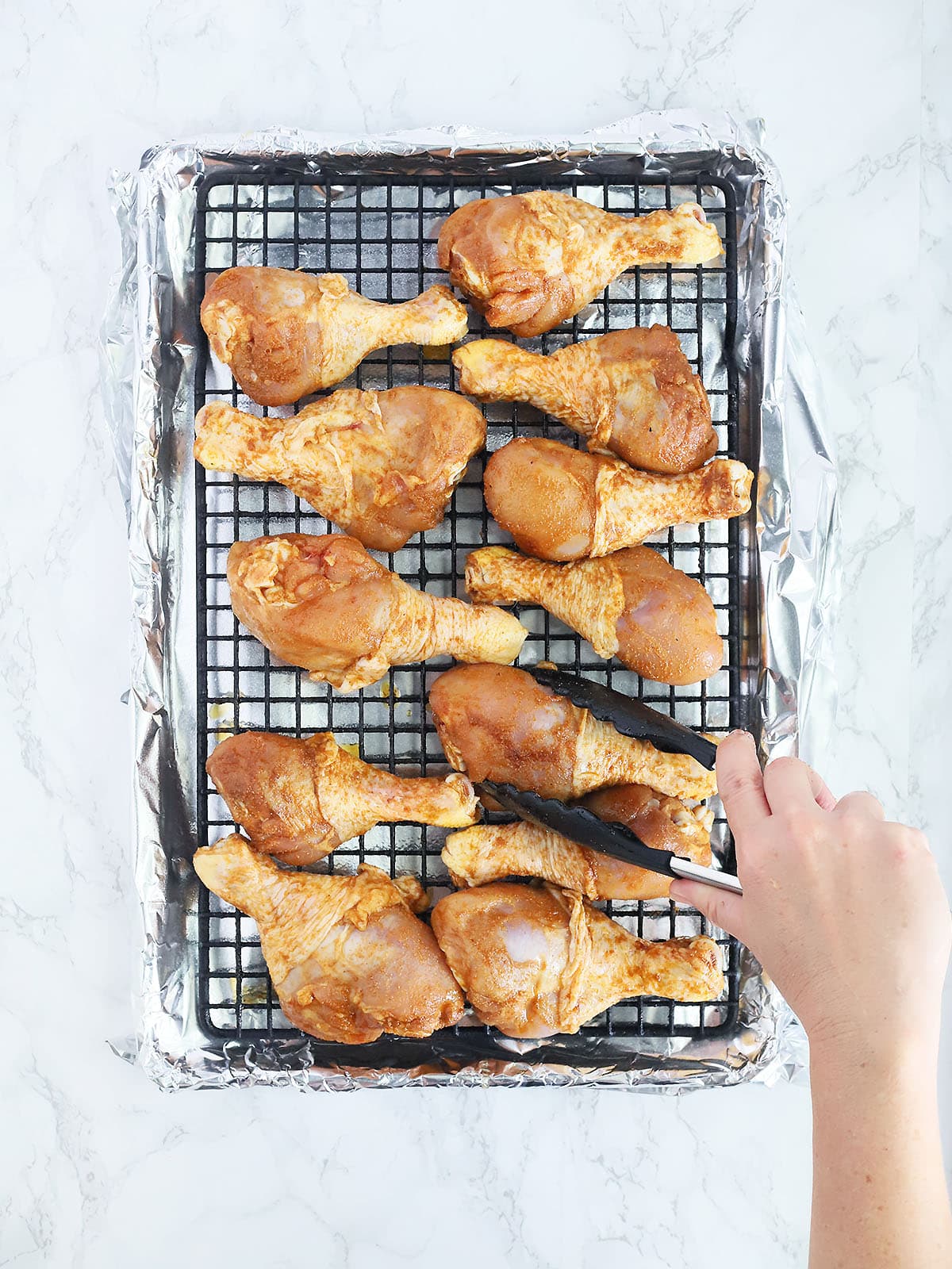 Using tongs to arrange uncooked chicken drumsticks on a baking sheet