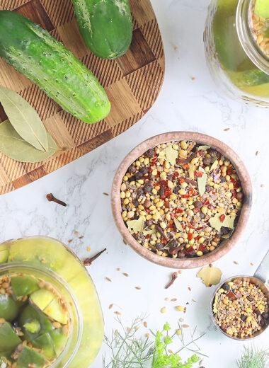 small clay bowl filled with homemade pickling spices with cucumbers and jars of pickles to the side