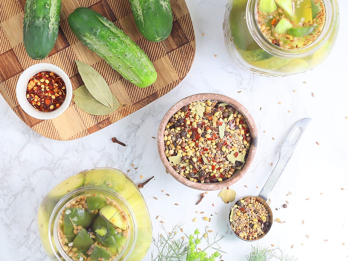 small clay bowl filled with homemade pickling spices with cucumbers and jars of pickles to the side