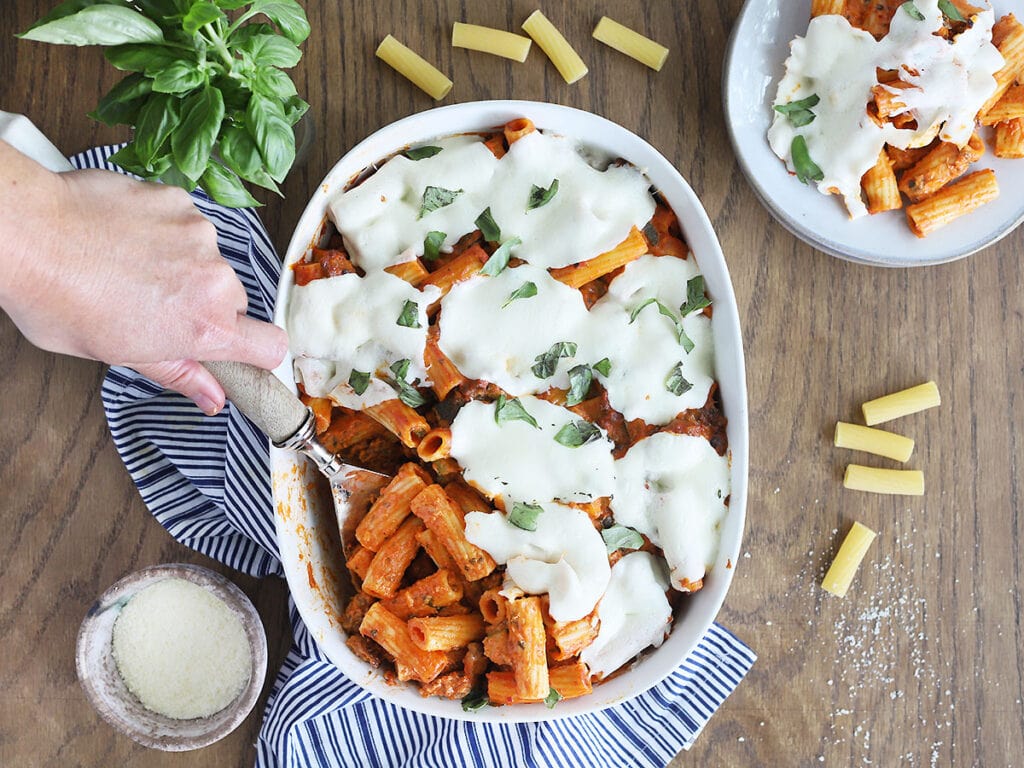 hand serving a portion of spicy rigatoni casserole from a white oval casserole dish