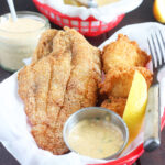 fried catfish fillet laying in a red basket with hushpuppies, remoulade sauce and lemon wedge