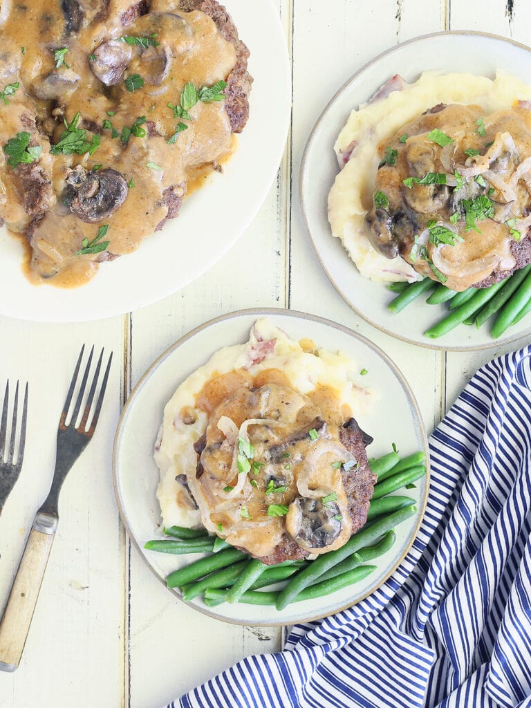 two white plates with hamburger steaks served over mashed potatoes and green beans with a serving plate of smothered hamburger steaks to the side
