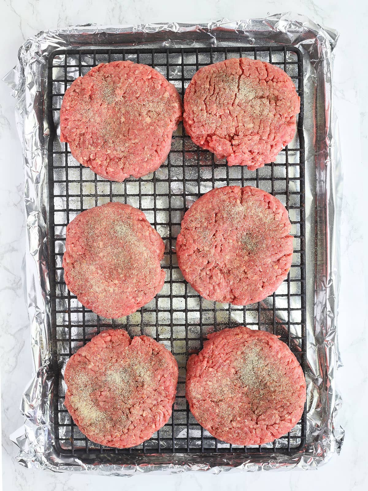 six seasoned uncooked hamburger patties on a baking rack placed over a foil lined baking sheet