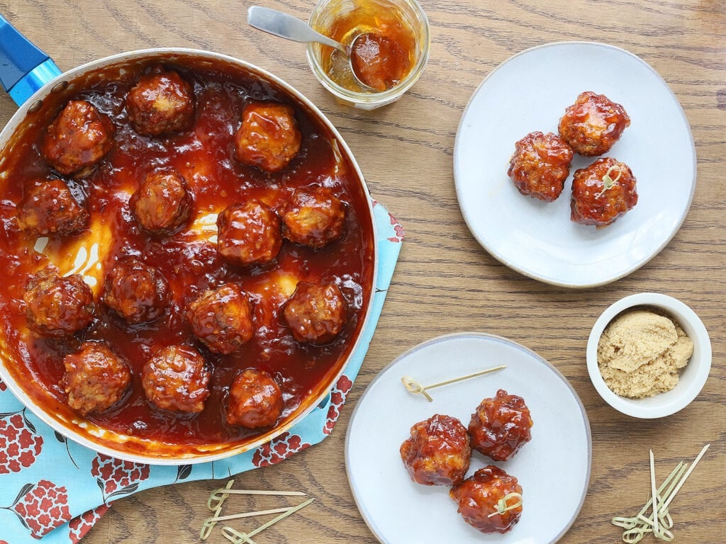 skillet of glazed meatballs with two plates containing three meatballs each to the side