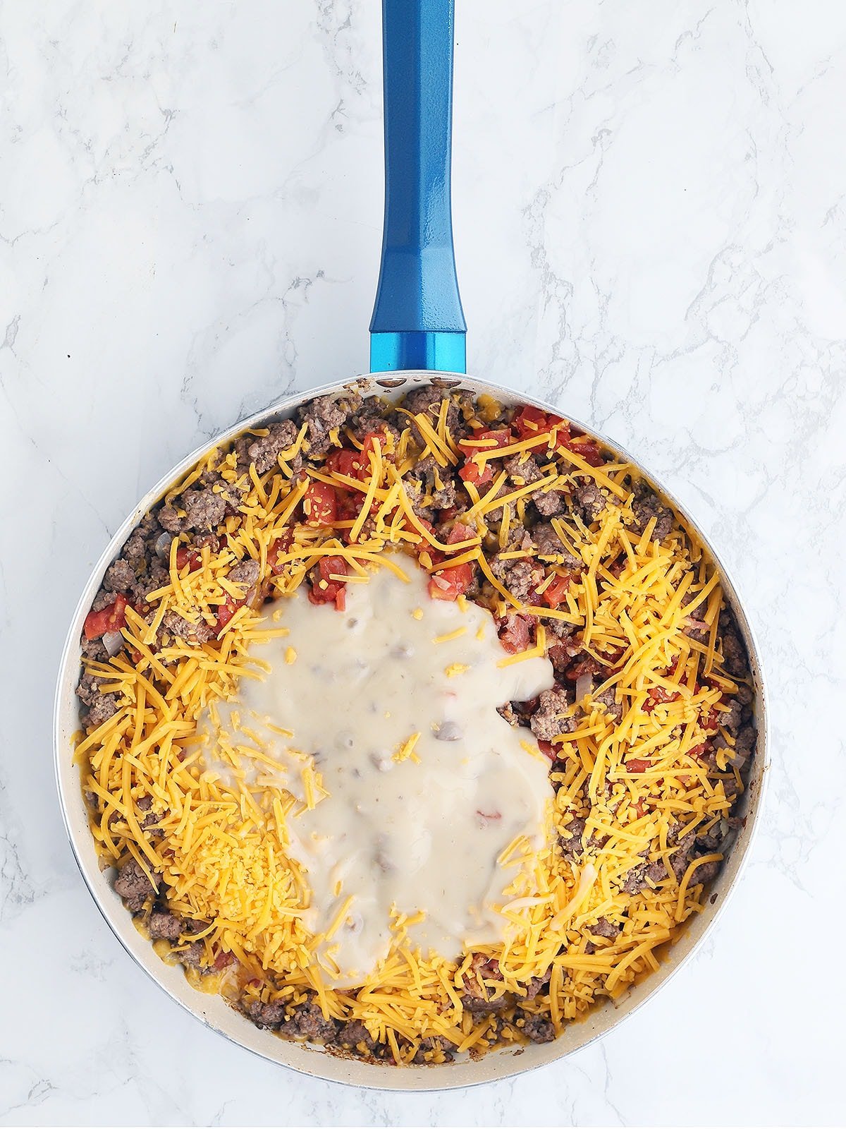 browned burger, tomatoes, soup mixture and shredded cheese in a blue skillet
