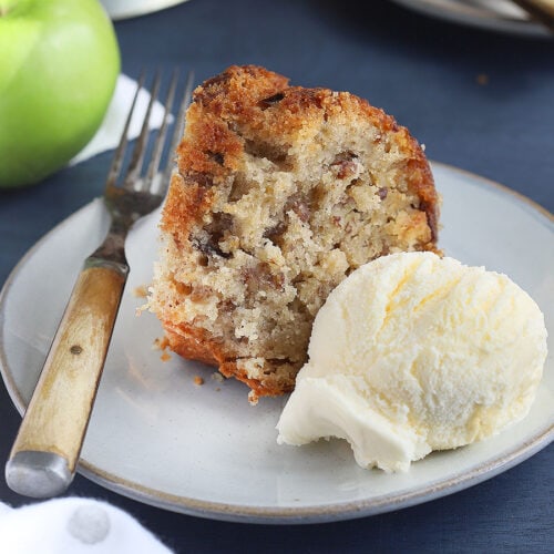 slice of apple dapple cake and ice cream on a white plate with a second slice of cake, a cake pedestal and green apple in the background