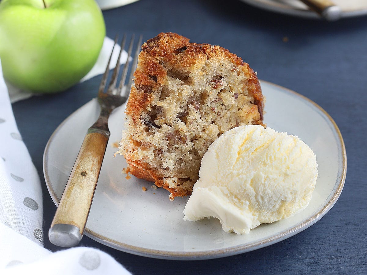 slice of apple dapple cake and ice cream on a white plate with a second slice of cake, a cake pedestal and green apple in the background