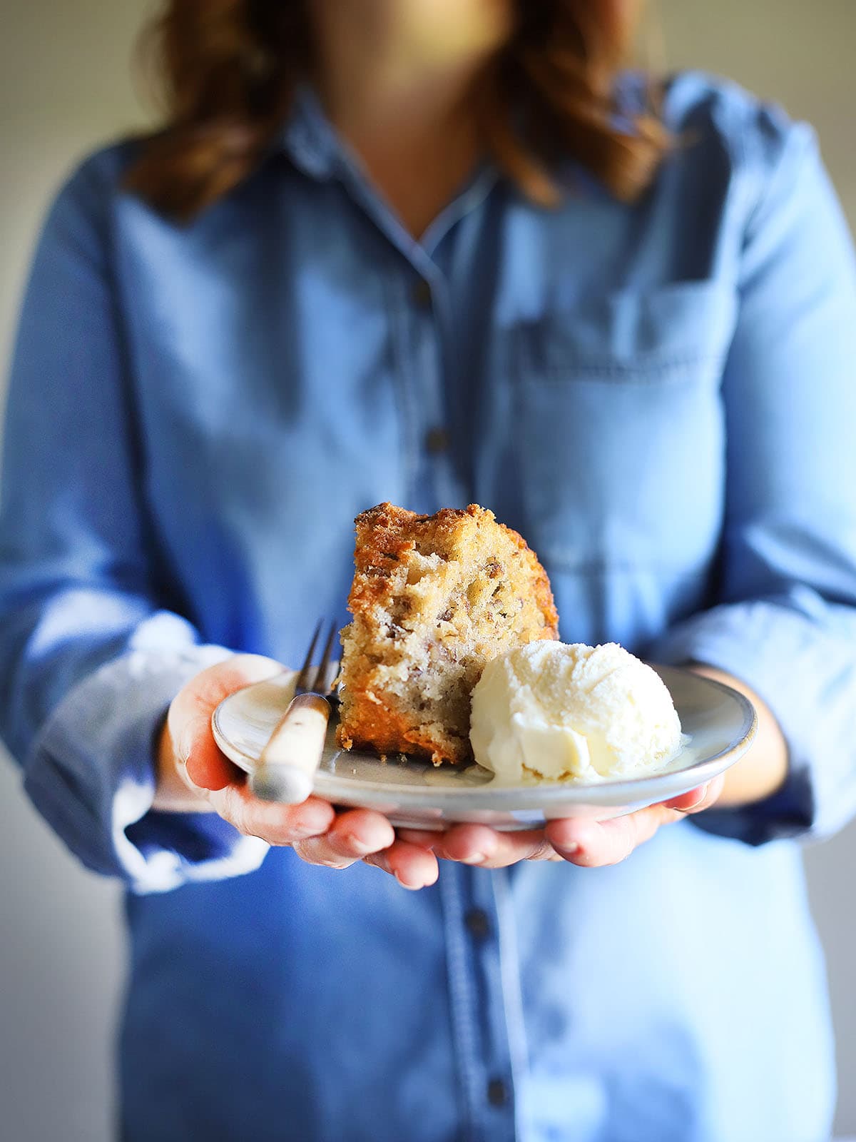 person in a denim shirt holding out a plate of apple dapple cake with ice cream