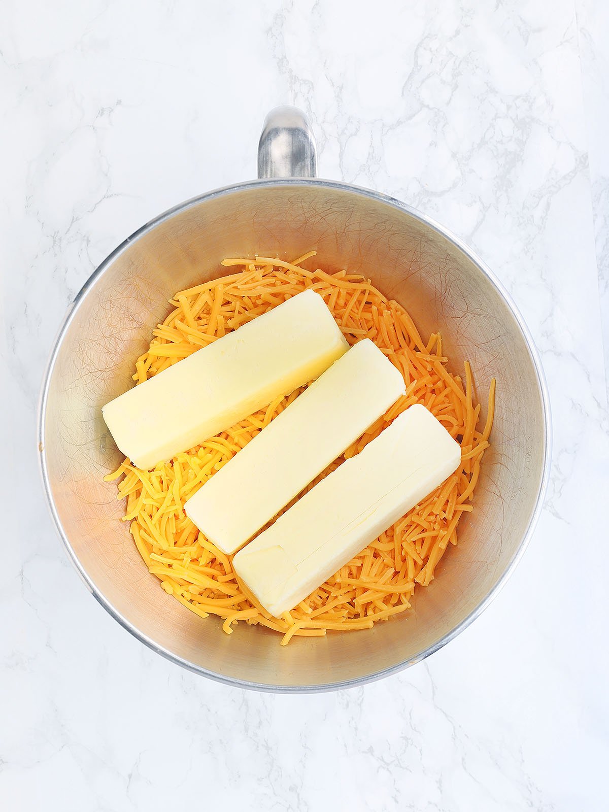 shredded cheddar cheese and three stick of butter in a metal mixing bowl.