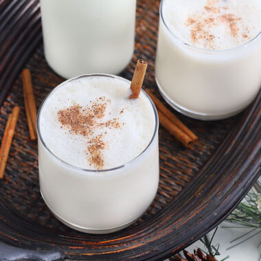 two glasses of bourbon milk punch garnished with nutmeg and cinnamon sticks on a wooden tray