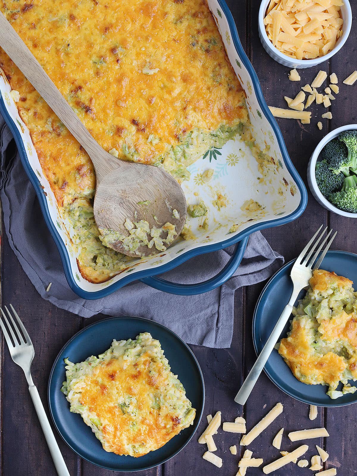 wooden serving spoon laid across the casserole dish with servings of cheesy broccoli casserole on the side