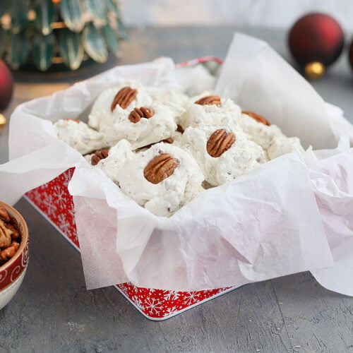 homemade divinity candy arranged in a paper lined candy tin with Christmas decorations in the background