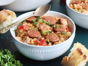 white bowl of chicken jambalaya with a pan, hot sauce and crusty bread in the background