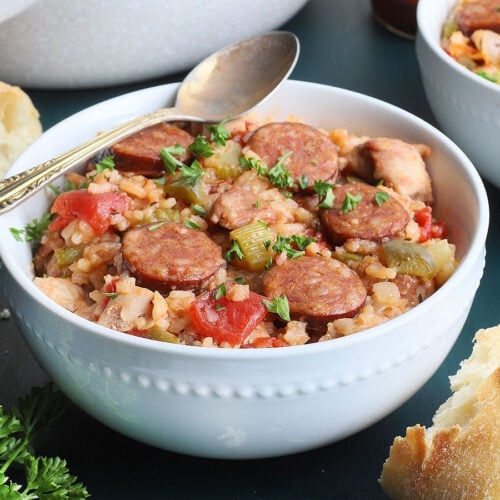 white bowl of chicken jambalaya with a pan, hot sauce and crusty bread in the background