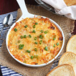 Dish of hot baked crawfish dip garnished with sliced green onions surrounded by toasted baguettes and a Mason jar of iced tea.