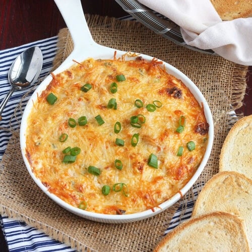 Dish of hot baked crawfish dip garnished with sliced green onions surrounded by toasted baguettes and a Mason jar of iced tea.