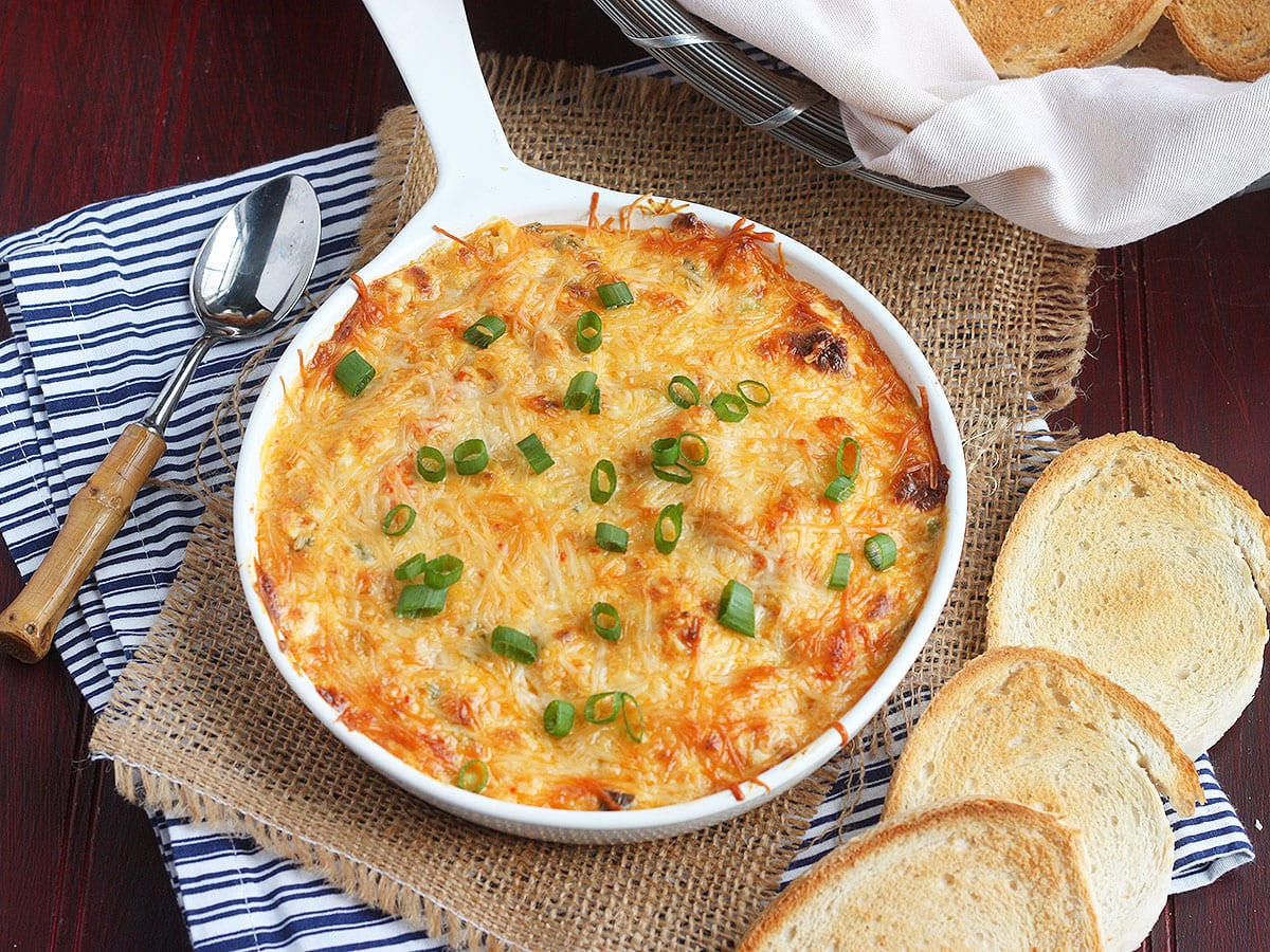 Dish of hot baked crawfish dip garnished with sliced green onions surrounded by toasted baguettes and a Mason jar of iced tea.