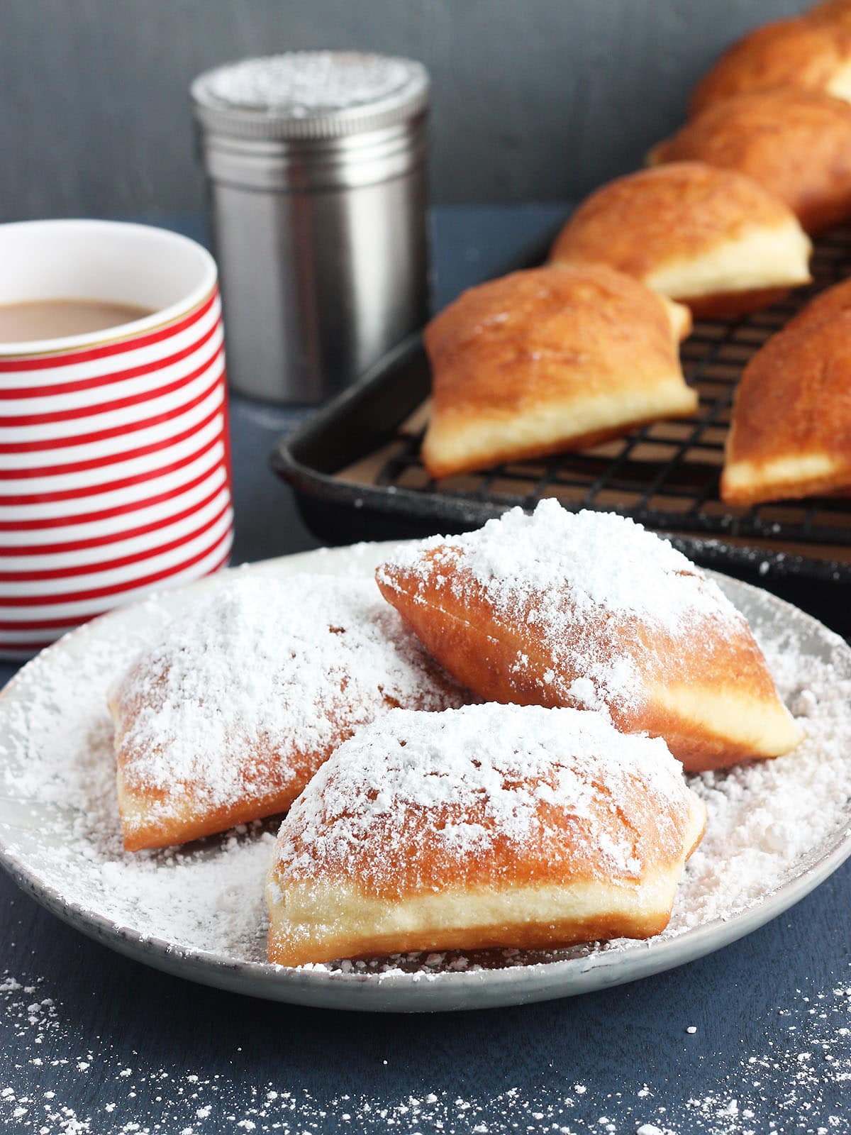 three beignets on a grey plate with powdered sugar with cafe au lait in the background