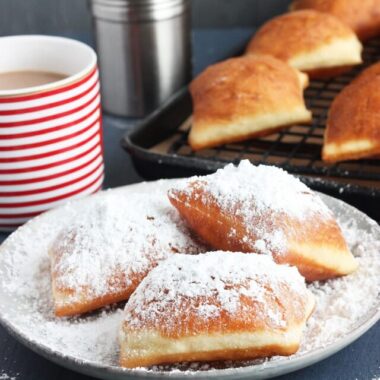 three beignets on a grey plate with powdered sugar with cafe au lait in the background