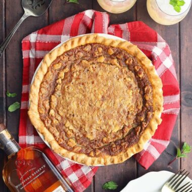Whole Kentucky Derby pie on a dark wooden background with plates, forks, two mint juleps and a bottle of bourbon to the side.