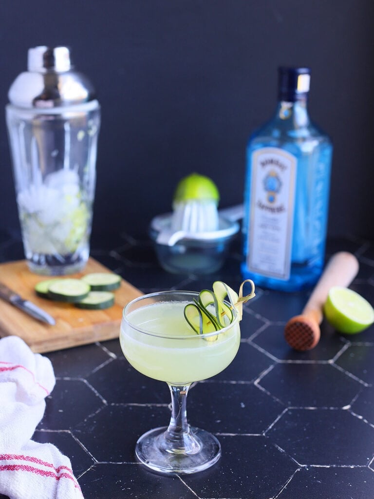 cucumber gimlet in a coupe glass garnished with a sliced cucumber and gimlet ingredients in the background.