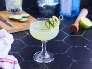 cucumber gimlet in a coupe glass garnished with a sliced cucumber and gimlet ingredients in the background.