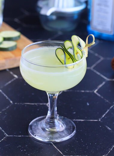 cucumber gimlet in a coupe glass garnished with a sliced cucumber and gimlet ingredients in the background.