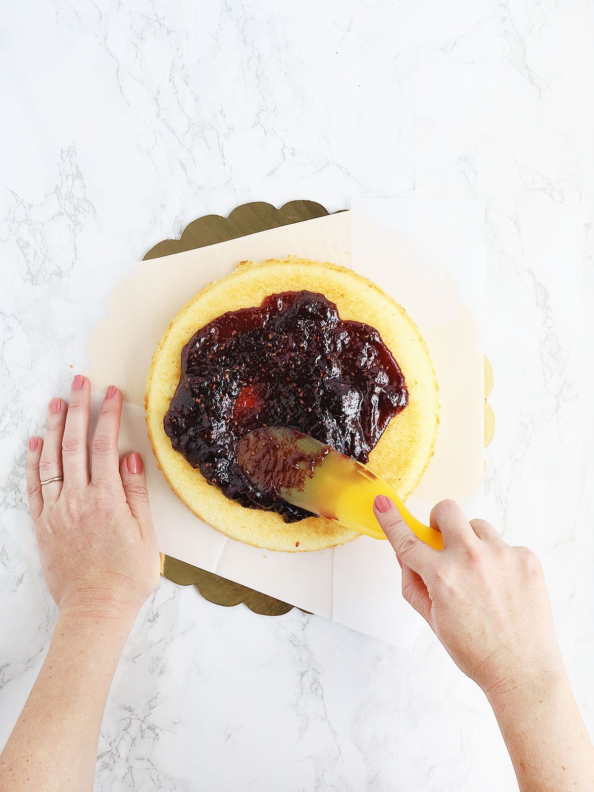 hands using a wide spatula to spread raspberry preserves over the top of one cake layer