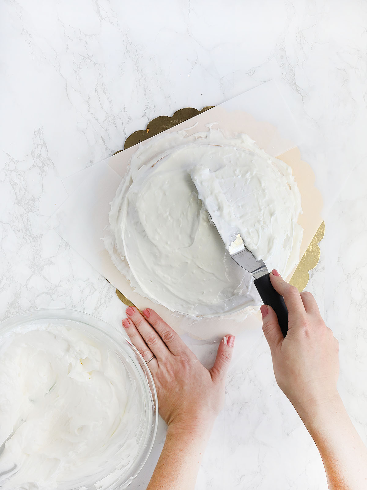 hand using an offset spatula with spread whipped cream icing over the top and the sides of the coconut cake