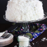 finished coconut cake on a glass cake pedestal with plates and a jar of coconut to the side.