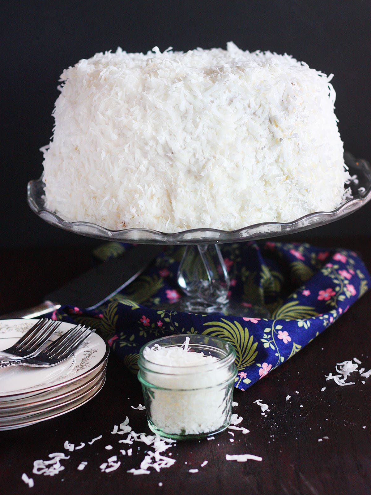 finished coconut cake on a glass cake pedestal with plates and a jar of coconut to the side.
