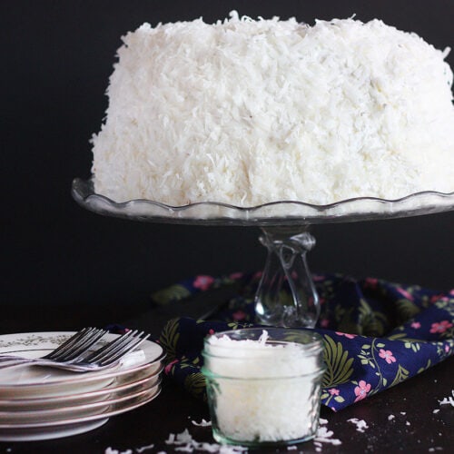 finished coconut cake on a glass cake pedestal with plates and a jar of coconut to the side.