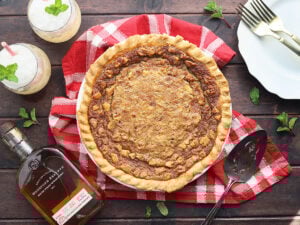 Whole Kentucky Derby pie on a dark wooden background with plates, forks, two mint juleps and a bottle of bourbon to the side.