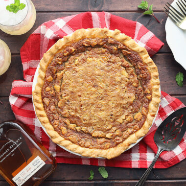 Whole Kentucky Derby pie on a dark wooden background with plates, forks, two mint juleps and a bottle of bourbon to the side.