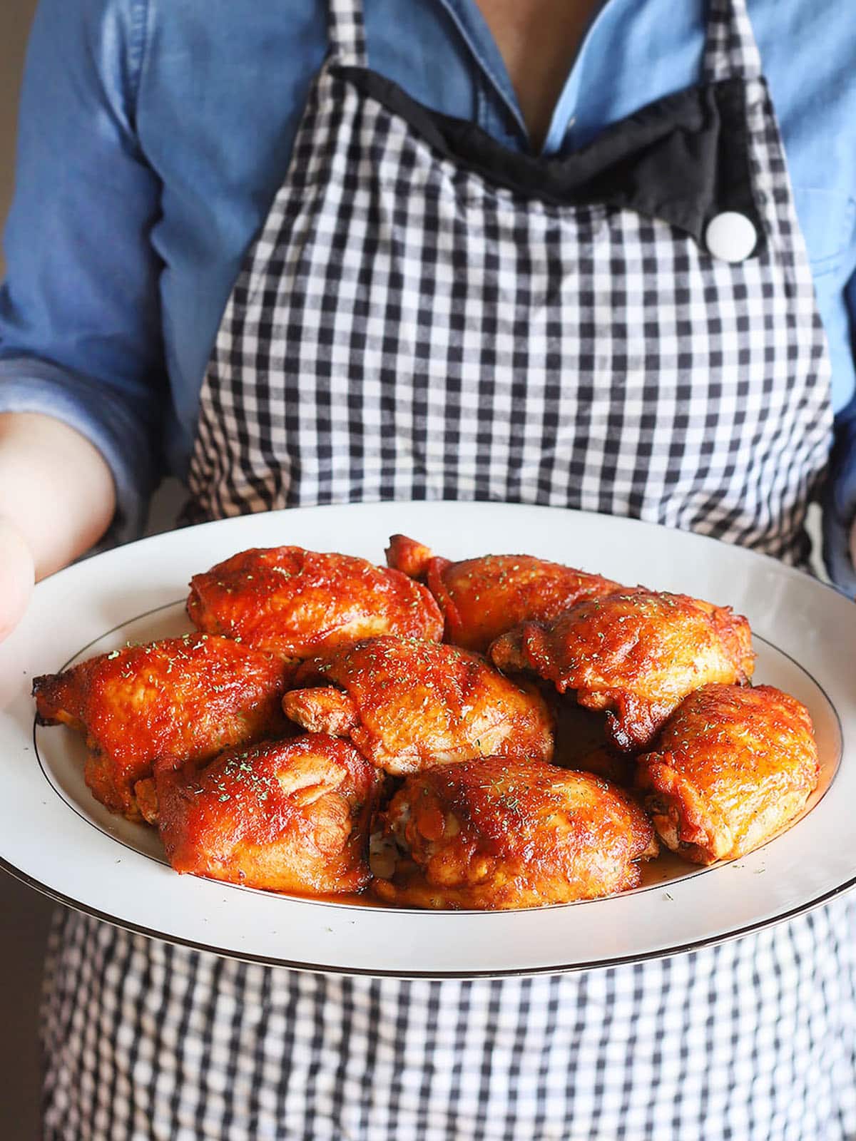 person in a black and white checked apron holding a white platter of oven baked BBQ chicken thighs.