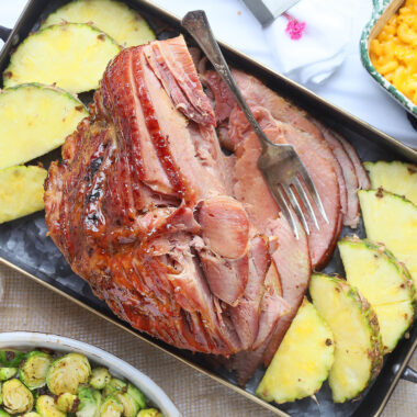 sliced pineapple glazed ham on a metal serving tray with pineapple slices, roasted Brussels sprouts and macaroni and cheese on the side