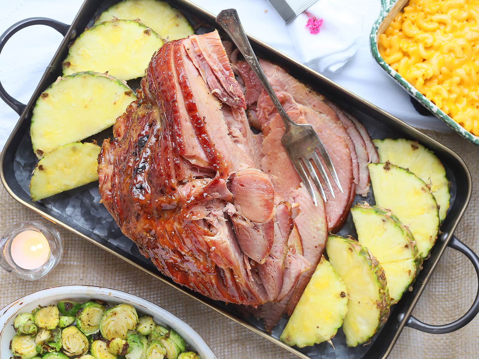 sliced pineapple glazed ham on a metal serving tray with pineapple slices, roasted Brussels sprouts and macaroni and cheese on the side