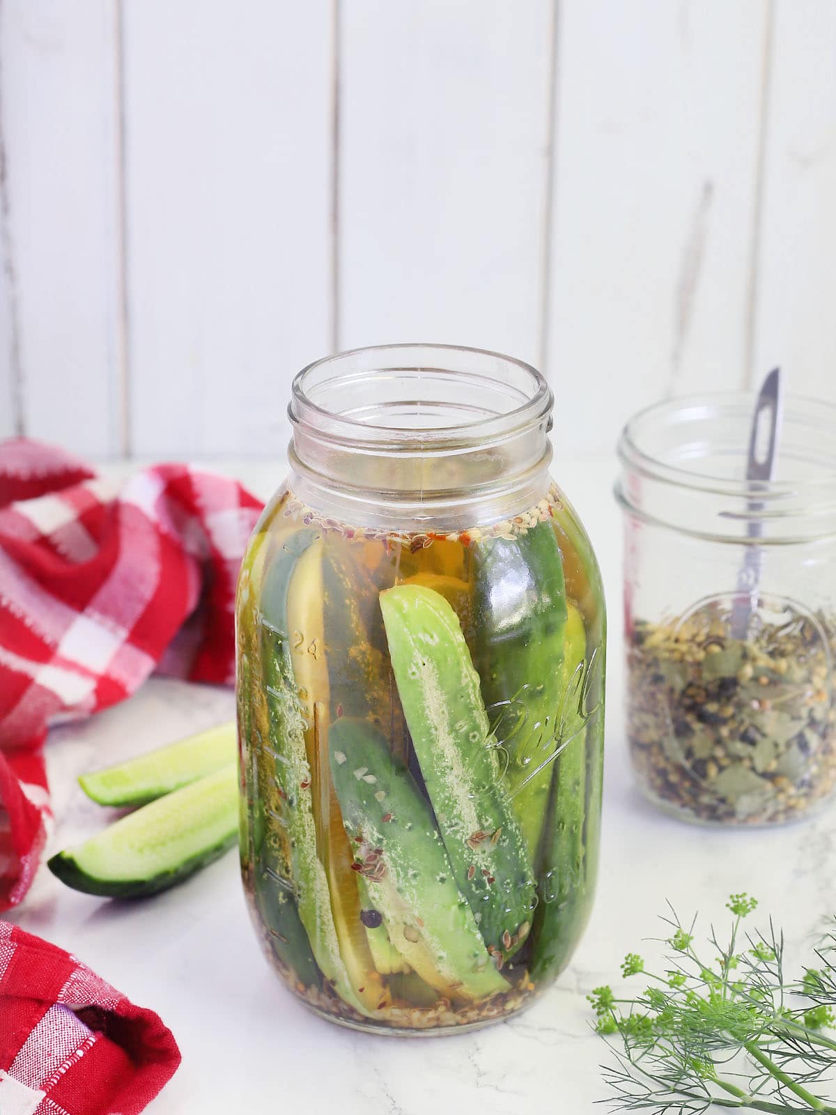 Quart mason jar of cucumbers spears in pickle brine with a jar of pickling spices and fresh cucumber spears in the background