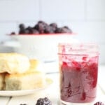 a jar of blackberry jam with a plate of biscuits and a colander of blackberries in the background