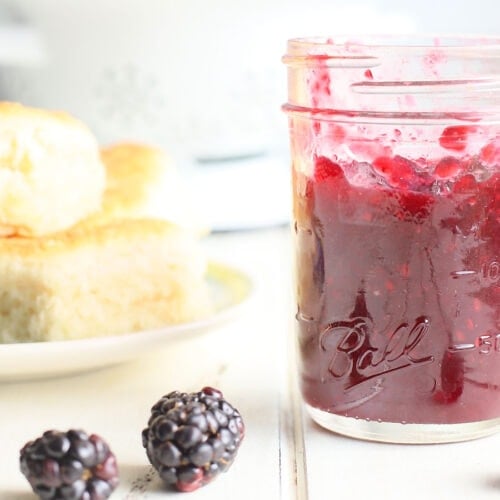 a jar of blackberry jam with a plate of biscuits in the background