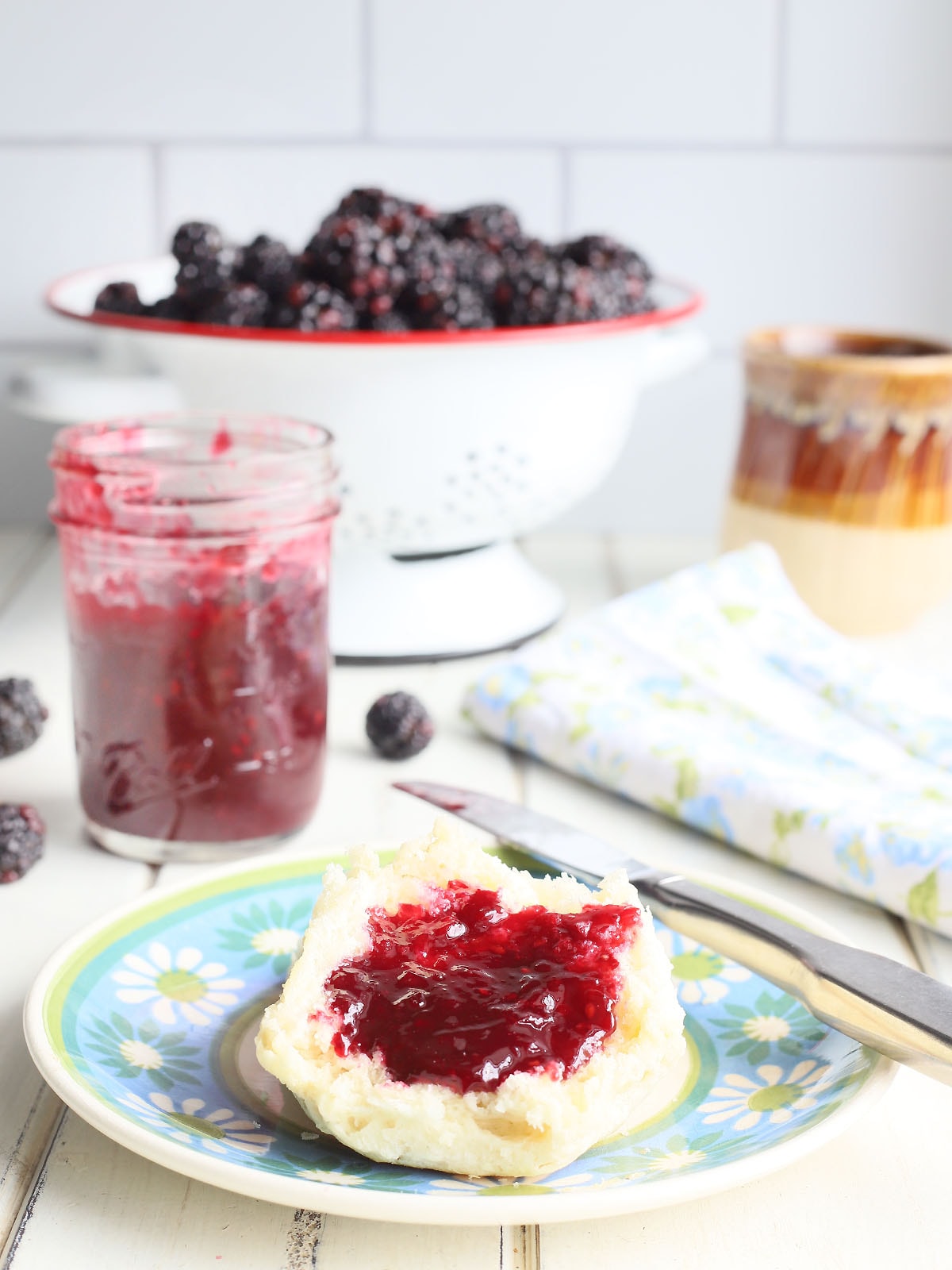 a blue floral plate with a biscuit covered in blackberry jam, a jar of jam, coffee cup and colander of whole berries in the background