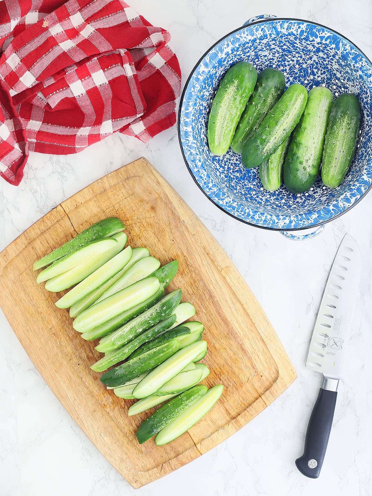 wooden cutting board with dill pickle spears, knife and blue colander with whole cucumbers on the side