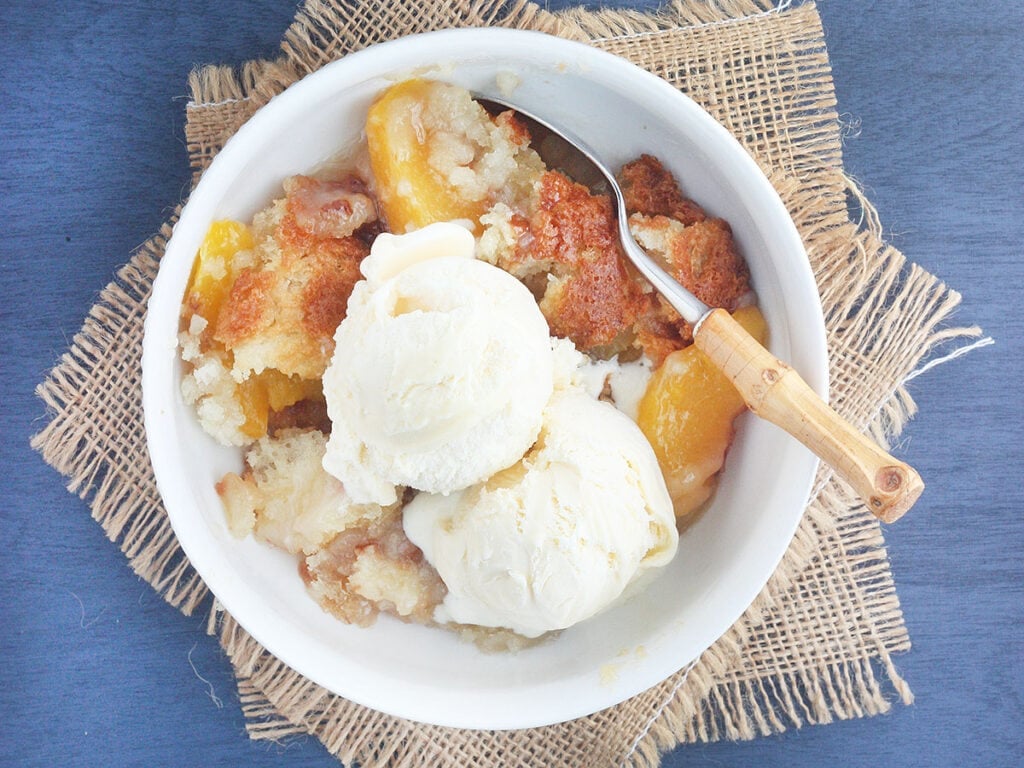 white bowl of peach cobbler topped with a scoop of vanilla ice cream