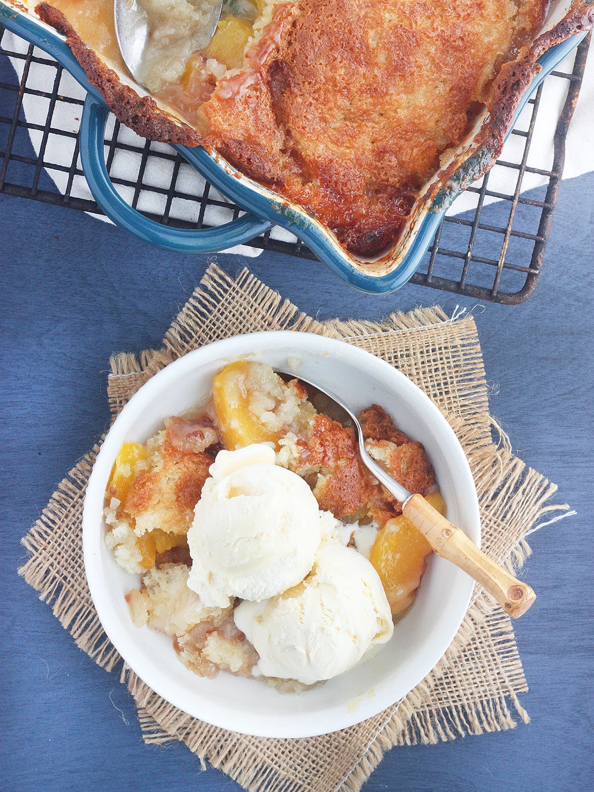 white bowl of peach cobbler topped with a scoop of vanilla ice cream with a pan of cobbler cooling on a rack to the side.