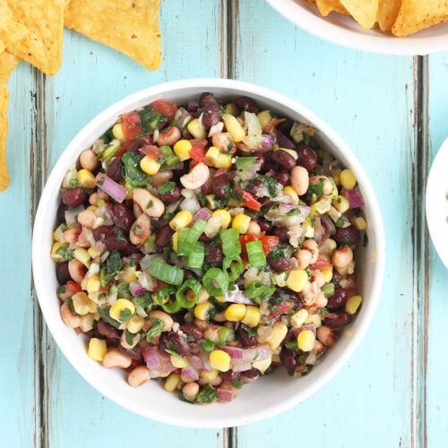 White bowl of Redneck Caviar on a blue background with a white bowl of tortilla chips and green onions on the side