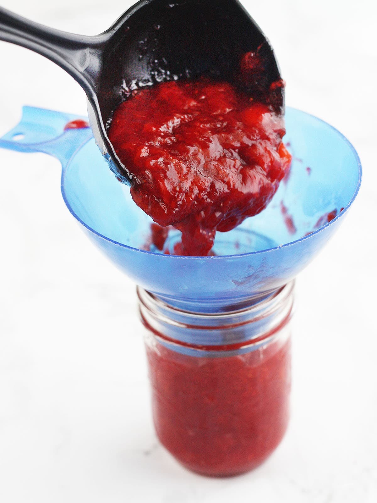 ladling strawberry jam into a mason jar with a blue wide mouth funnel