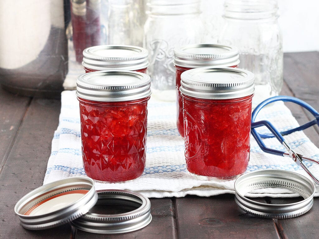 four jars of homemade jam cooling on a towel with empty mason jars and canning rings in the background.